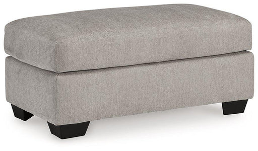 A beige Avenal Park Ottoman by Ashley Furniture with a fabric upholstery and wooden block legs, placed on a hardwood floor.